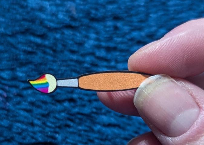 Hand holding a paintbrush with a rainbow design on a blue textured background, displayed in an app interface.