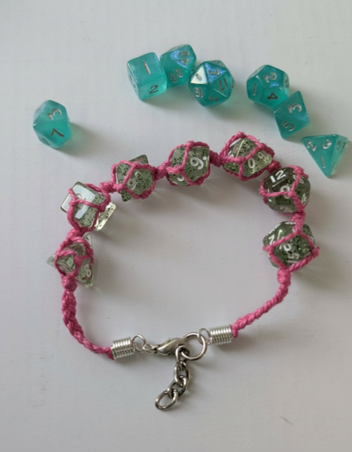 A pink braided cord bracelet with a lobster clasp, glittery translucent polyhedral dice, translucent teal polyhedral gaming dice are also scattered around on a plain light background.