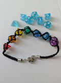 A handmade braided cord bracelet with colorful translucent polyhedral dice beads in red, orange, yellow, green, blue, and purple, shown with matching translucent blue polyhedral dice on a white background.