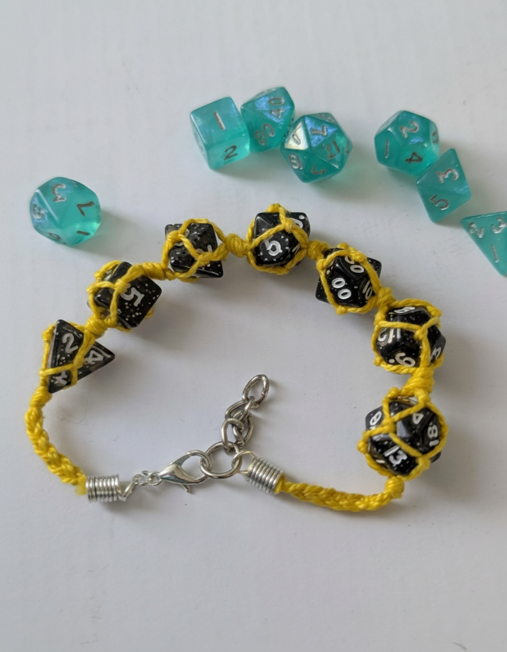 A yellow braided cord bracelet with black polyhedral gaming dice printed with white numbers, shown on a white background with several translucent teal dice scattered above.