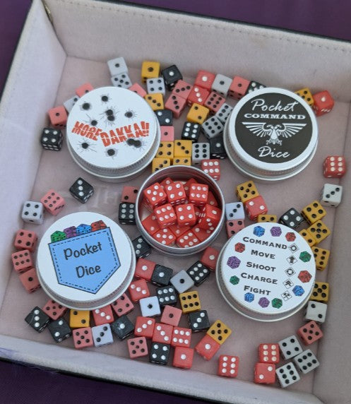 In a dice tray are a set of miniature gaming dice and four labeled metal tins arranged inside. The tins read 'More Dakka!!', 'Pocket Command Dice', 'Pocket Dice', and 'Command,  Move, Shoot, Charge, Fight', with many small red, black, white, yellow, and multicolored dice scattered around.