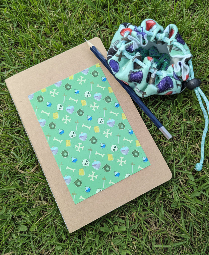A flat-lay of a kraft paper notebook with a green patterned cover, a blue pencil, and a mint green drawstring pouch, all arranged on grass.