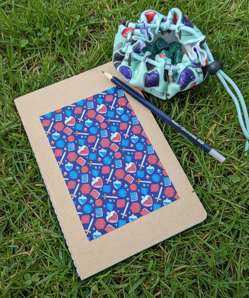 A flat-lay of a kraft paper notebook with a blue and red patterned cover, a blue pencil, and a mint green drawstring pouch all arranged together on grass
