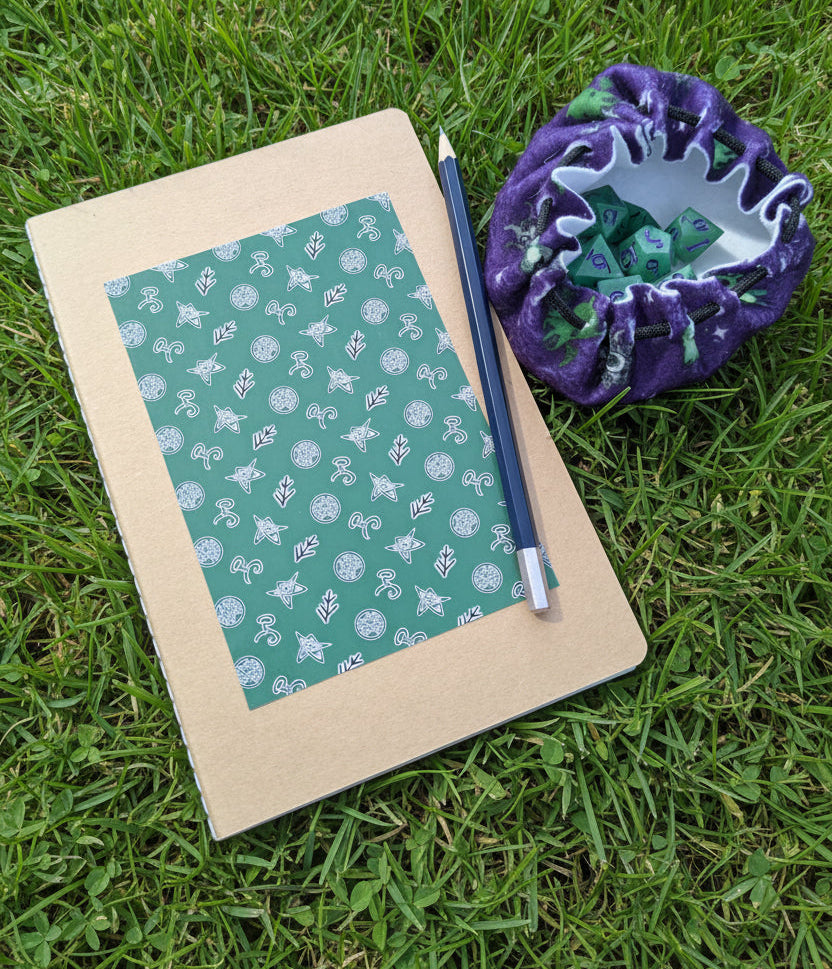 A kraft paper notebook with a green patterned cover lies on grass, next to a blue pencil and a purple drawstring bag filled with green dice.