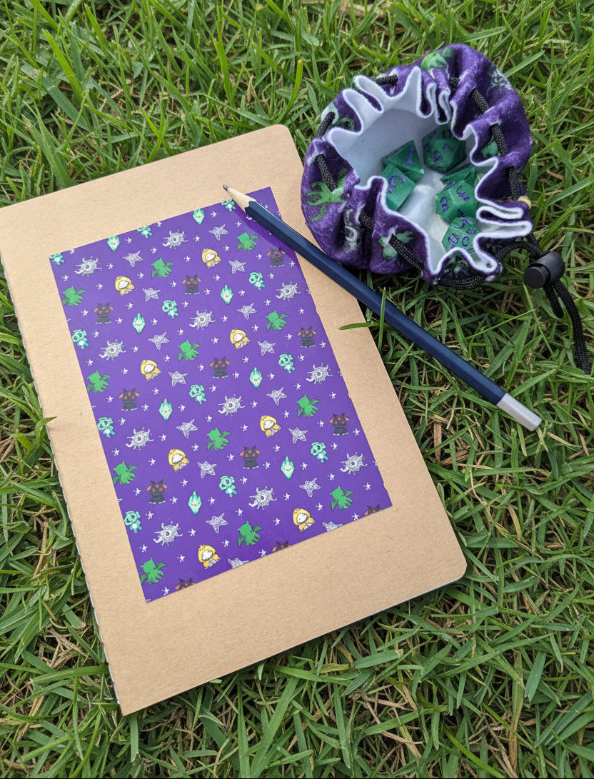 A flat-lay of a kraft paper notebook with a purple patterned cover, a blue pencil, and a purple and green patterned drawstring pouch all arranged on grass.