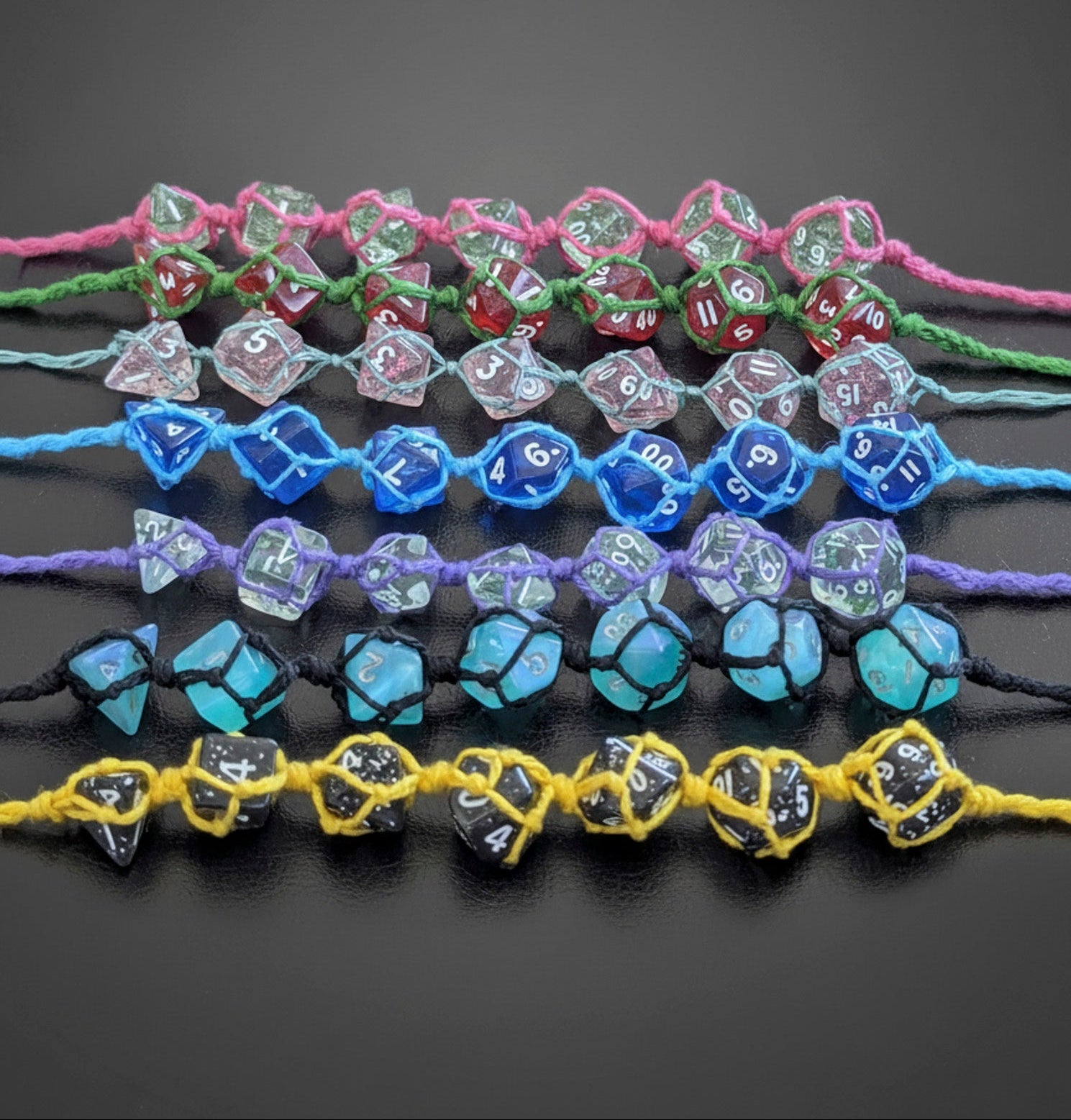 Seven sets of translucent polyhedral gaming dice made into macrame  bracelets arranged horizontally on a dark gray background. The sets include pink, green, light blue, purple, black, and yellow cords, with dice in clear, blue, green, red and black.
