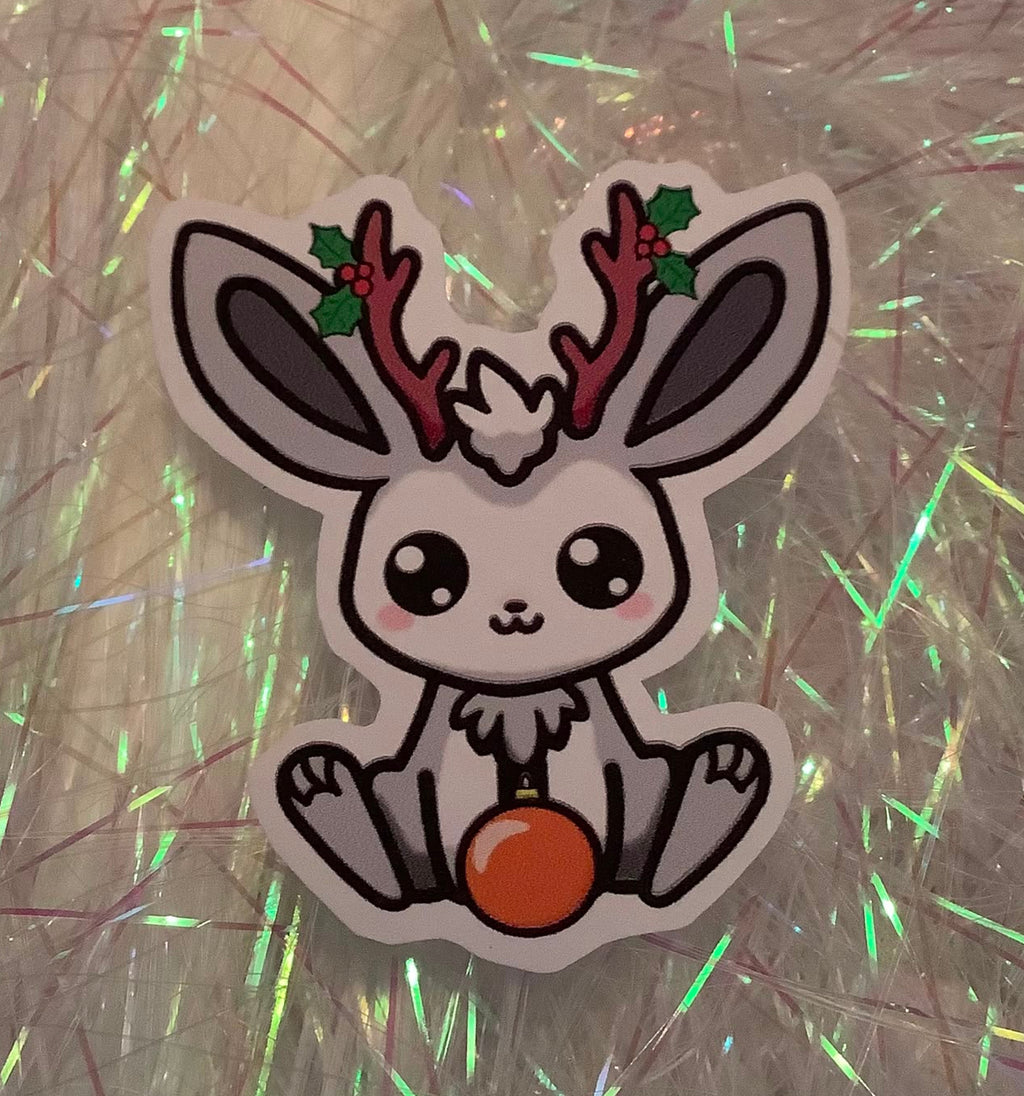 A die-cut sticker featuring a cute cartoon jackalope, green holly leaves, and a orange ornament, displayed on a sparkly silver background.