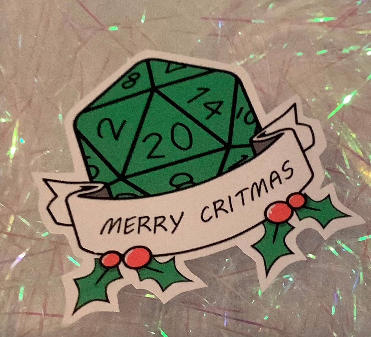 A die-cut sticker featuring a green 20-sided die with black numbers, a white banner reading 'MERRY CRITMAS', and green holly leaves with red berries, placed on a shiny silver foil background.