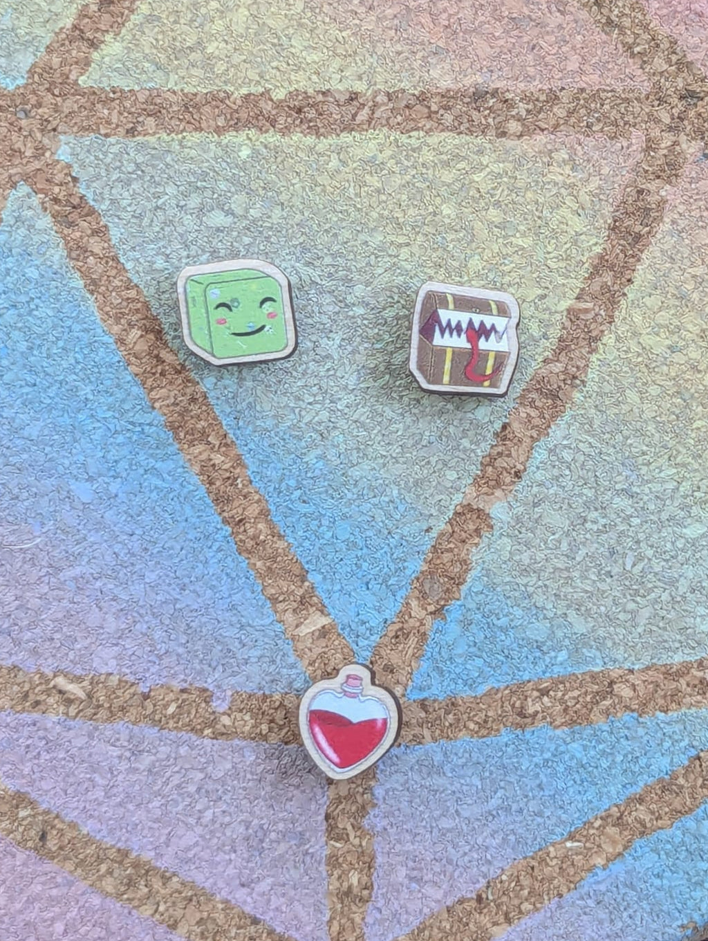 Three small wooden pins displayed on a round display board with geometric lines. The pins include a gelatinous smiling cube, a mimic chest and red  healing potion.