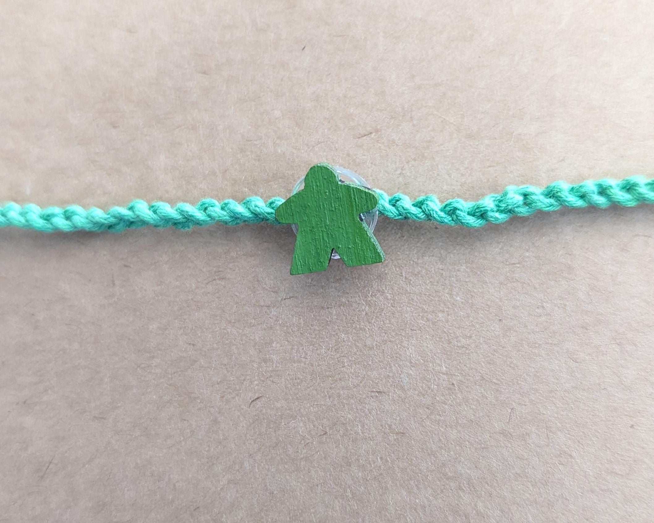 Green meeple charm on a plain green braided cord against a beige background