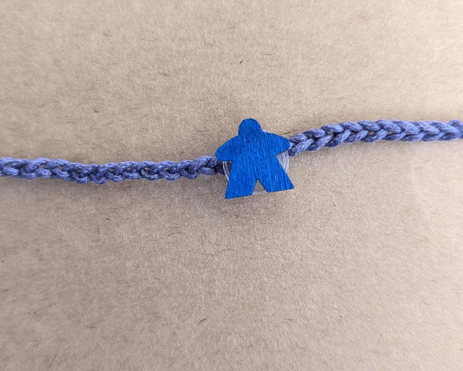 Blue braided bracelet and blue meeple charm on a beige background