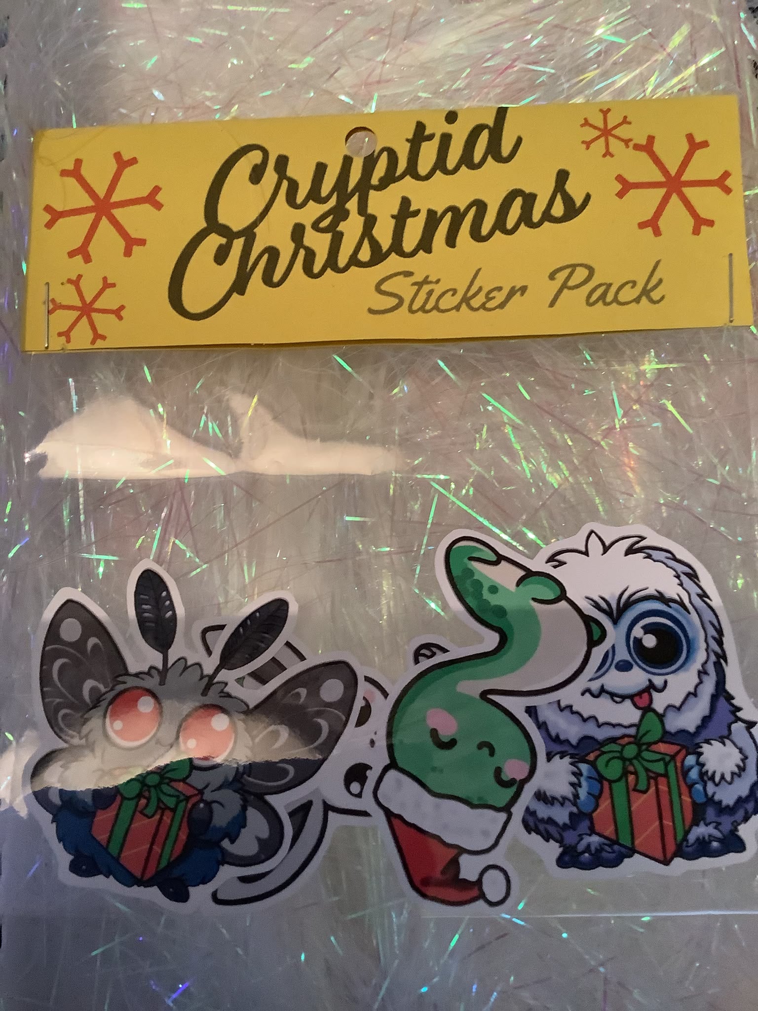 A Christmas sticker pack in clear plastic wrapping, featuring a yellow package label that reads 'Cryptid Christmas Sticker Pack' with red snowflakes, and two cartoon-style stickers.