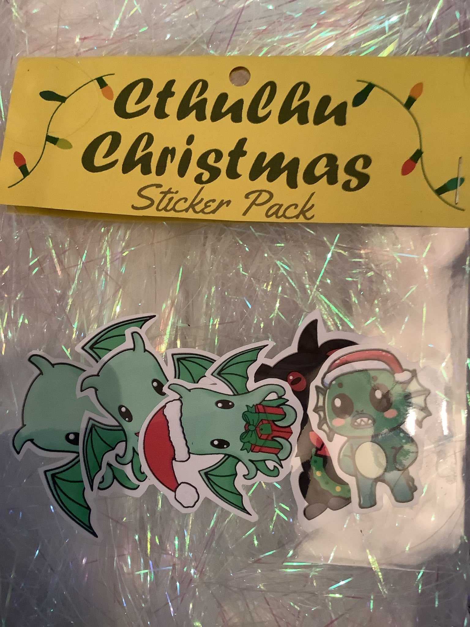 A Christmas sticker pack featuring elder gods cthulhu wearing festive holiday accessories, sealed in clear plastic packaging next to a yellow paper label that reads 'Cthulhu Christmas Sticker Pack'.