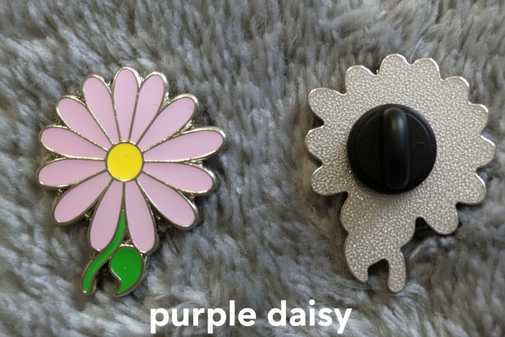 
A close-up of a flower-shaped enamel pin shown from both sides on a fuzzy gray surface. The front shows a purple daisy flower with a yellow center and green stem, and the back shows a black clasp on a silver scalloped metal base.
