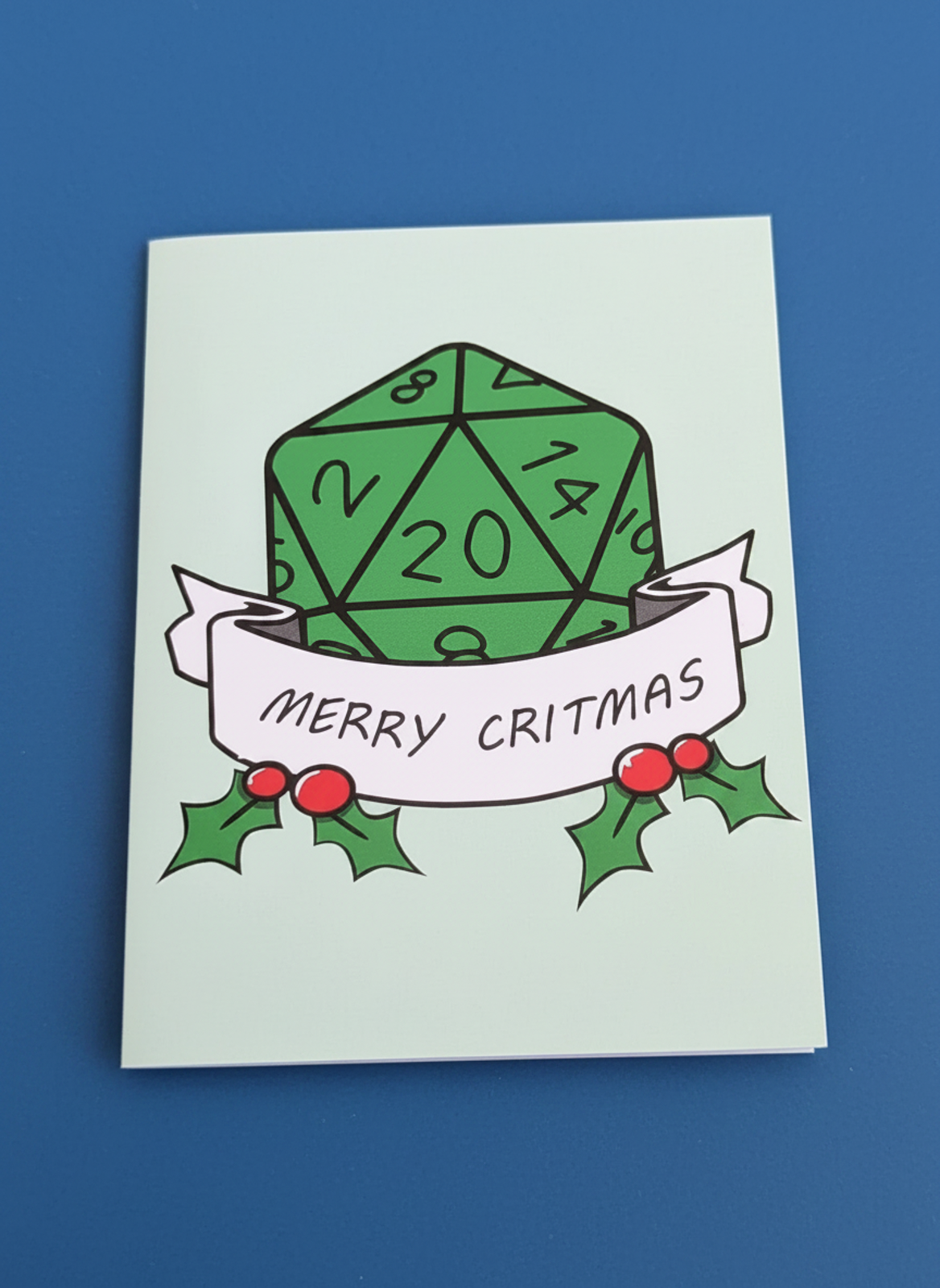 A greeting card featuring a green 20-sided die with 'MERRY CRITMAS' on a white banner, decorated with holly leaves and red berries, set against a solid blue background.
