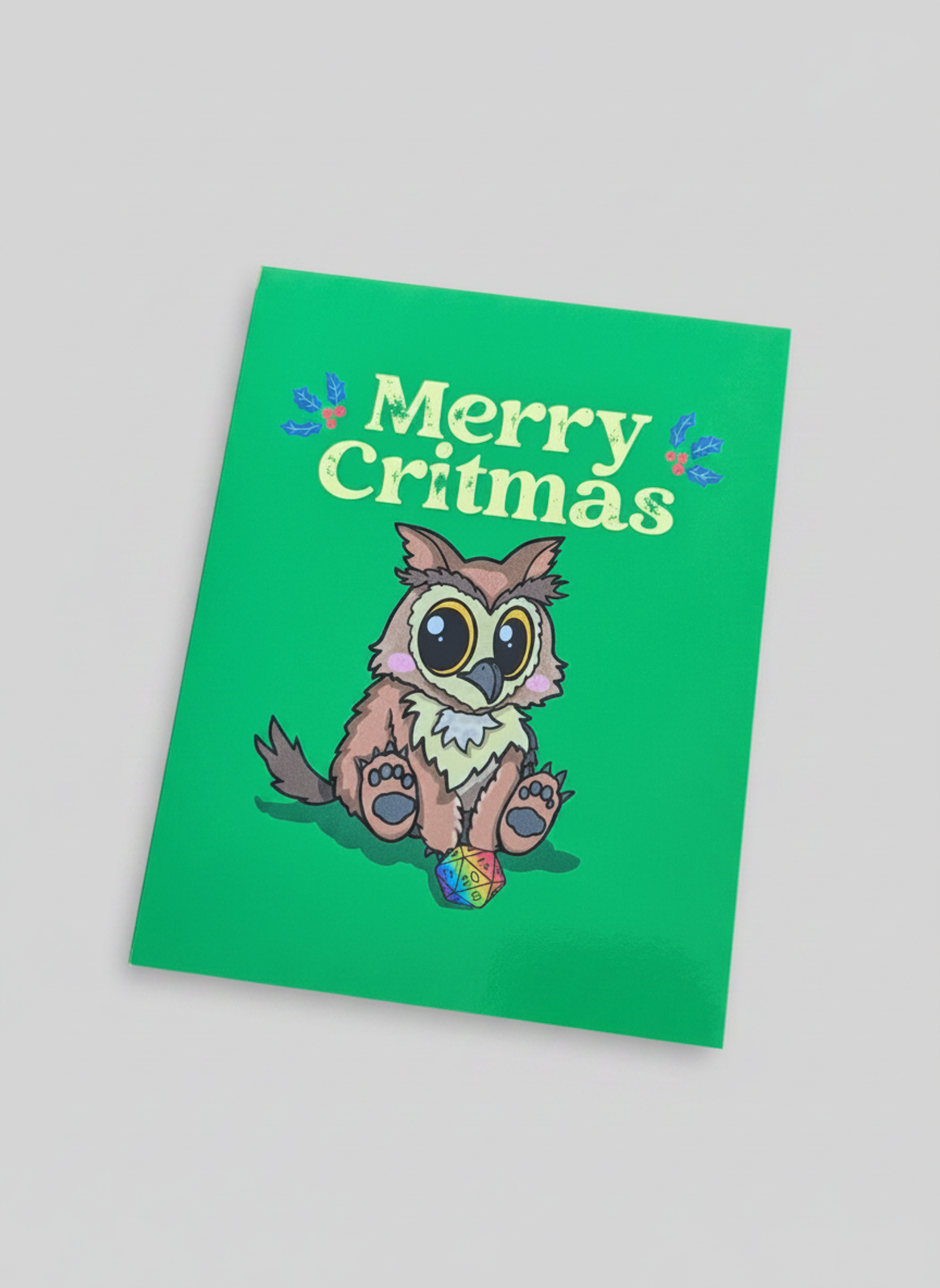 A green greeting card featuring a cute cartoon owlbear and the text 'Merry Critmas' in a light yellow-green font, shown at an angle on a neutral gray background.