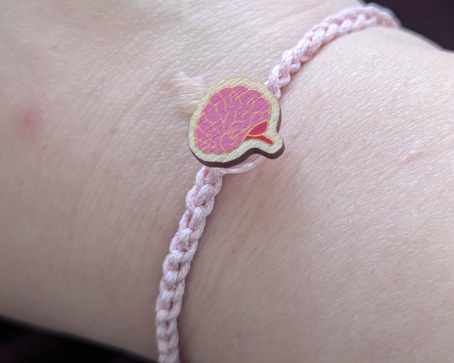 A close-up of a person's wrist wearing a light pink braided cord bracelet with a small wooden brain charm.