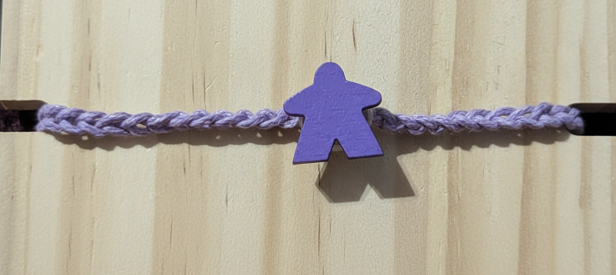 A light purple braided bracelet with a purple wooden meeple charm, displayed on a light wood background.