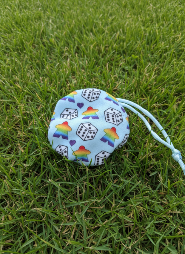 A small light blue drawstring pouch with a repeating pattern of white dice, rainbow stars, and purple hearts, resting on a green grass lawn.
