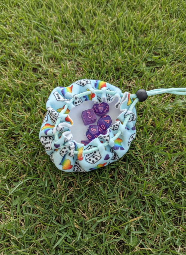 A small drawstring pouch made from light blue fabric with a black-and-white dice pattern and rainbow accents, placed on green grass. The pouch is open and contains purple polyhedral dice.