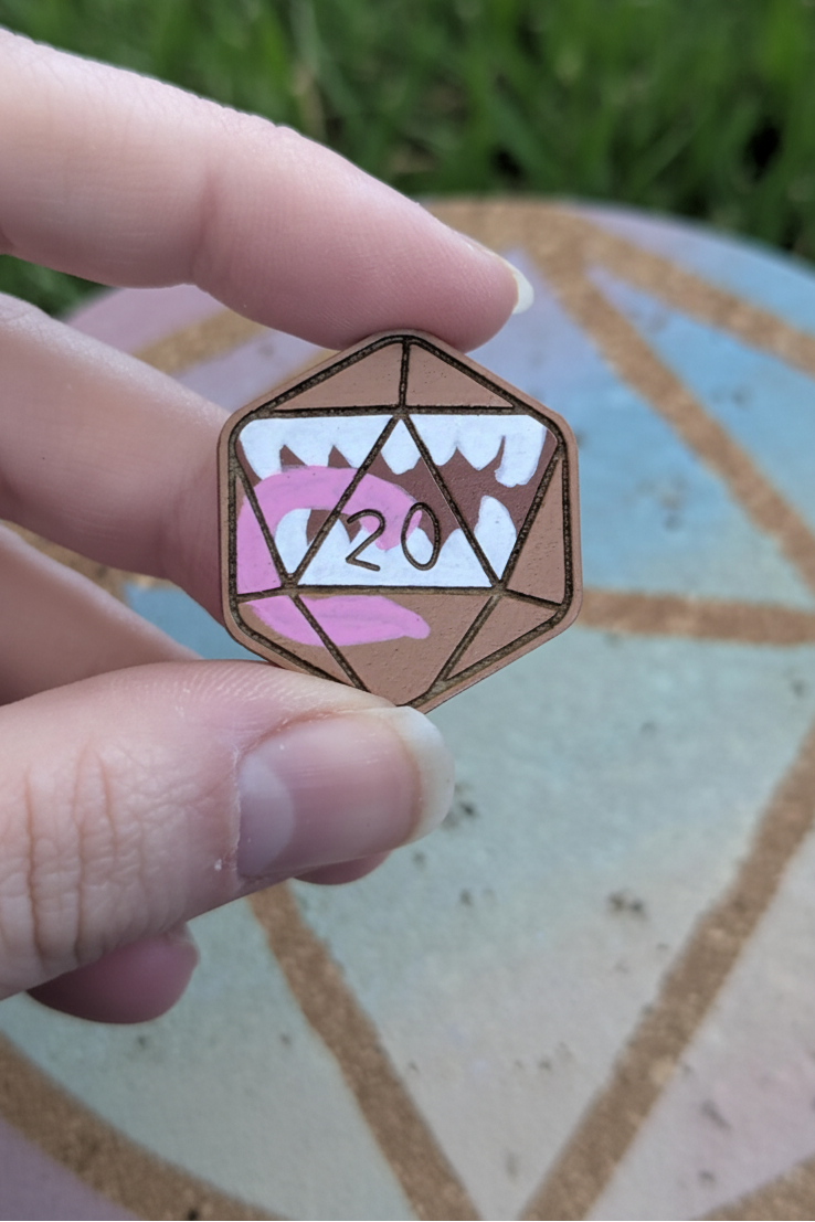 A hand holding a small mimic pin shaped like a 20-sided die, with a pink tongue and white teeth against a blurred grass background.