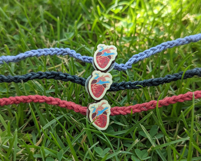 Three braided cord bracelets in light blue, black, and coral red, each featuring a small anatomical heart charm, laid out on a grass background.