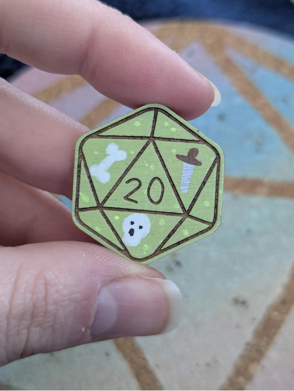 A hand holding a small die-cut pin shaped like a green 20-sided d20 decorated with white bone, skull, and dagger illustrations.