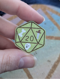 A hand holding a small die-cut pin shaped like a green 20-sided d20 decorated with white bone, skull, and dagger illustrations.