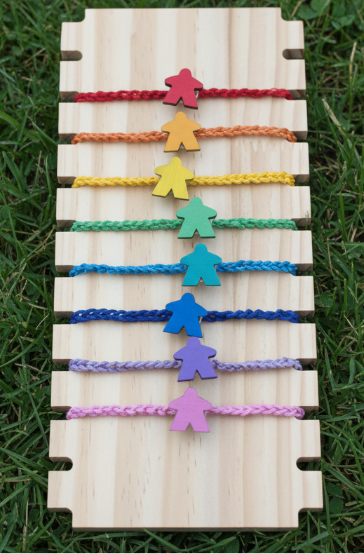 Nine colorful braided cord bracelets with matching wooden meeple charms are displayed on a light wood board, set against a grass background.