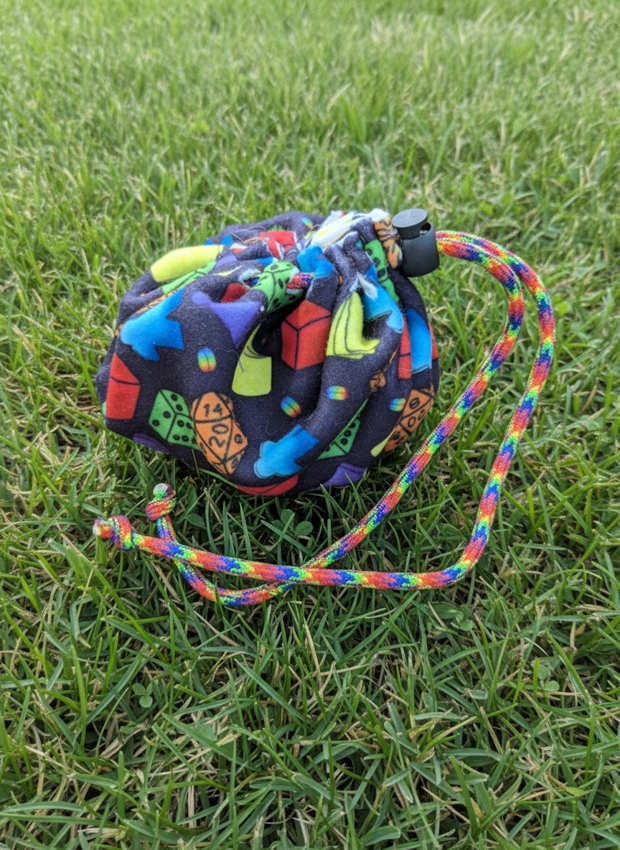 A small drawstring pouch with a colorful geometric pattern, resting on green grass. The pouch has a black plastic toggle closure and a braided drawstring strap with rainbow colors.