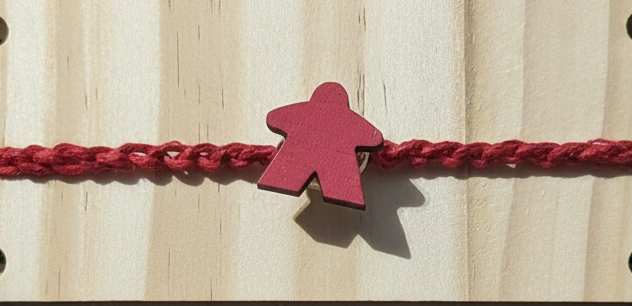 A red braided cord bracelet with a small red wooden meeple charm, shown against a light wood background.
