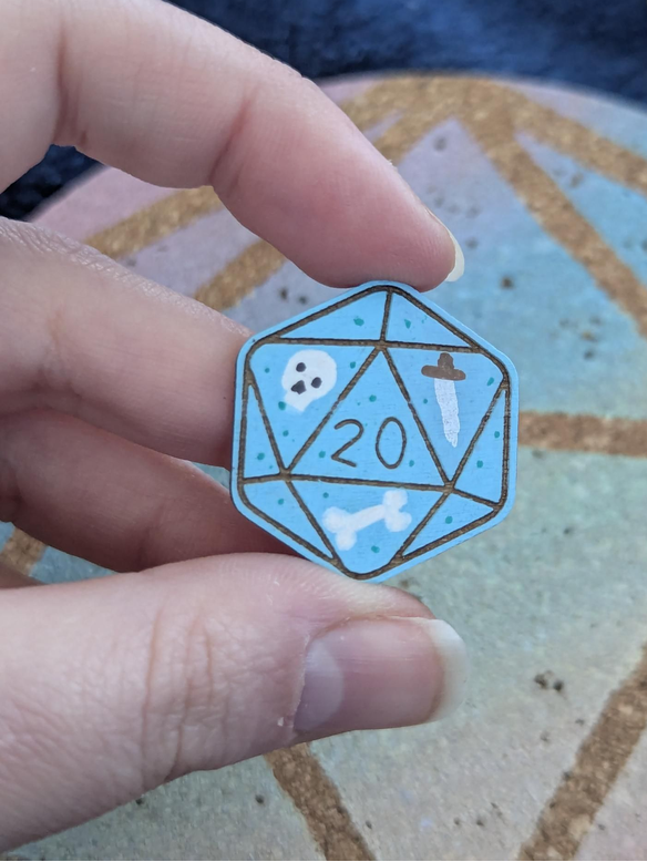 A hand holding a light blue d20 (twenty-sided die) pin painted blue with skull, crossbones, and a dagger inside.