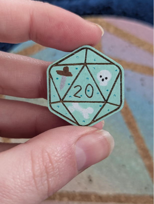 A close-up of a hand holding a die-cut wooden pin shaped like a 20-sided d20 light mint green in colour with a white skull, and white bones inside.