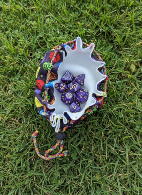 A small drawstring pouch with a colorful, patterned exterior and a white interior bowl, placed on green grass. The pouch contains several purple polyhedral gaming dice.