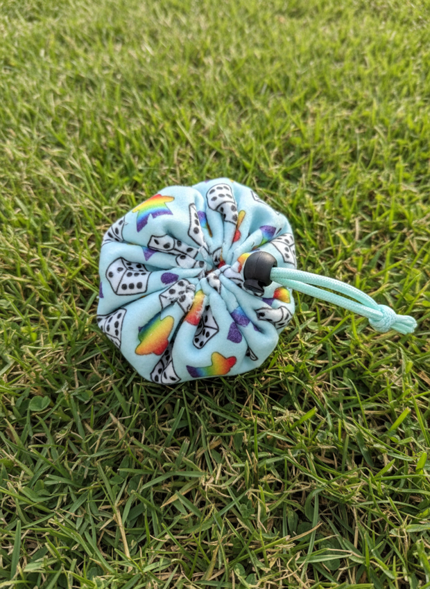 A small drawstring pouch made of light blue fabric with a colorful dice-and meeples print, resting on green grass.