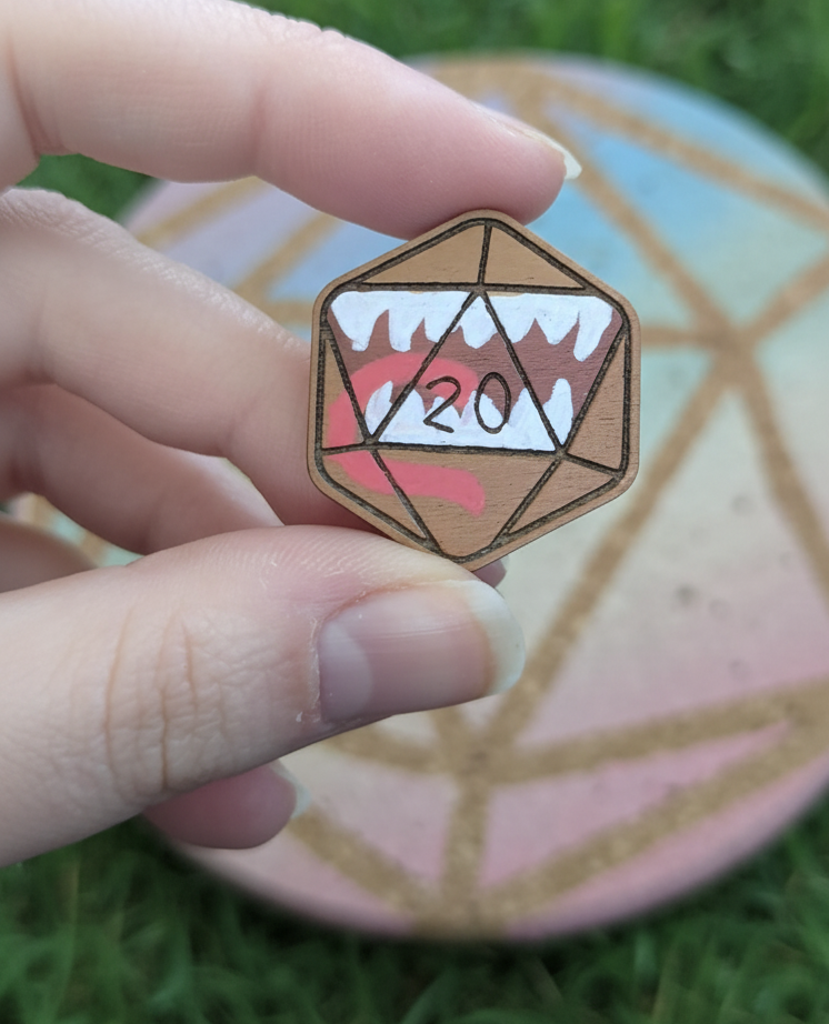 A hand holding a small pin shaped like a 20-sided die, with wood-grain texture brown body, white teeth, red tongue.