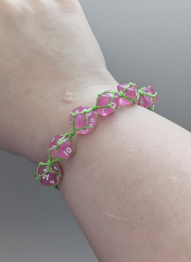 A close-up of a person's wrist wearing a handmade bracelet made of translucent pink polyhedral dice linked together with bright green cord.