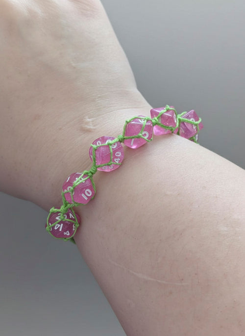 A close-up of a person's wrist wearing a handmade bracelet made of translucent pink polyhedral dice linked together with bright green cord.