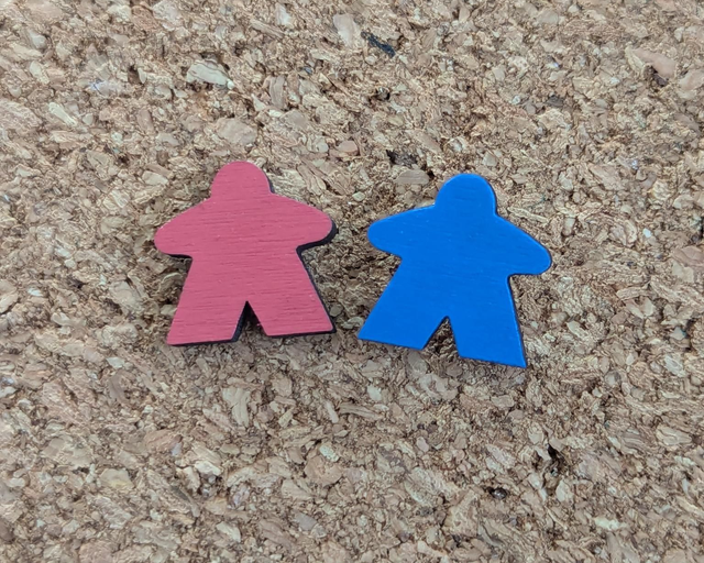 Two small wooden meeple pins, one red and one blue, placed side-by-side on a brown cork board background.