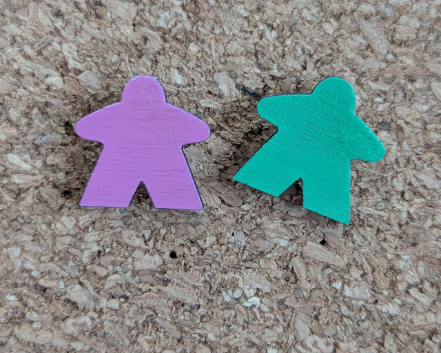 Two small wooden game pieces meeples one painted purple and the other green, placed on a cork surface.