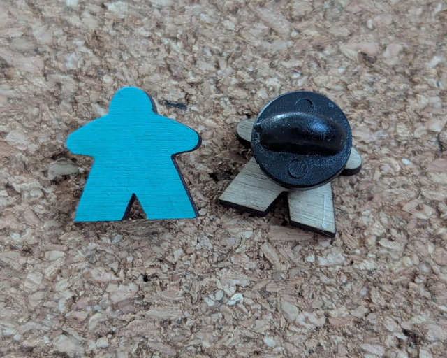 A turquoise blue wooden game piece meeple stands next to one turned round to show the pin and rubber clasp on a cork board background.
