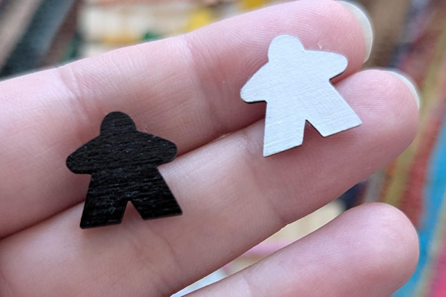 Two small wooden meeple pins, one black and one white, held on a person's fingers.