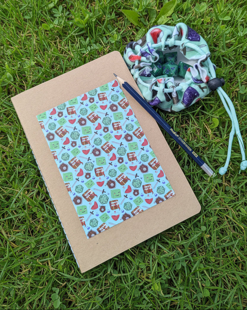 A handmade fabric drawstring pouch in a mint green and purple patterned material, shown next to a kraft paper notebook with a colorful patterned paper front and a blue pencil, all laid out on grass.