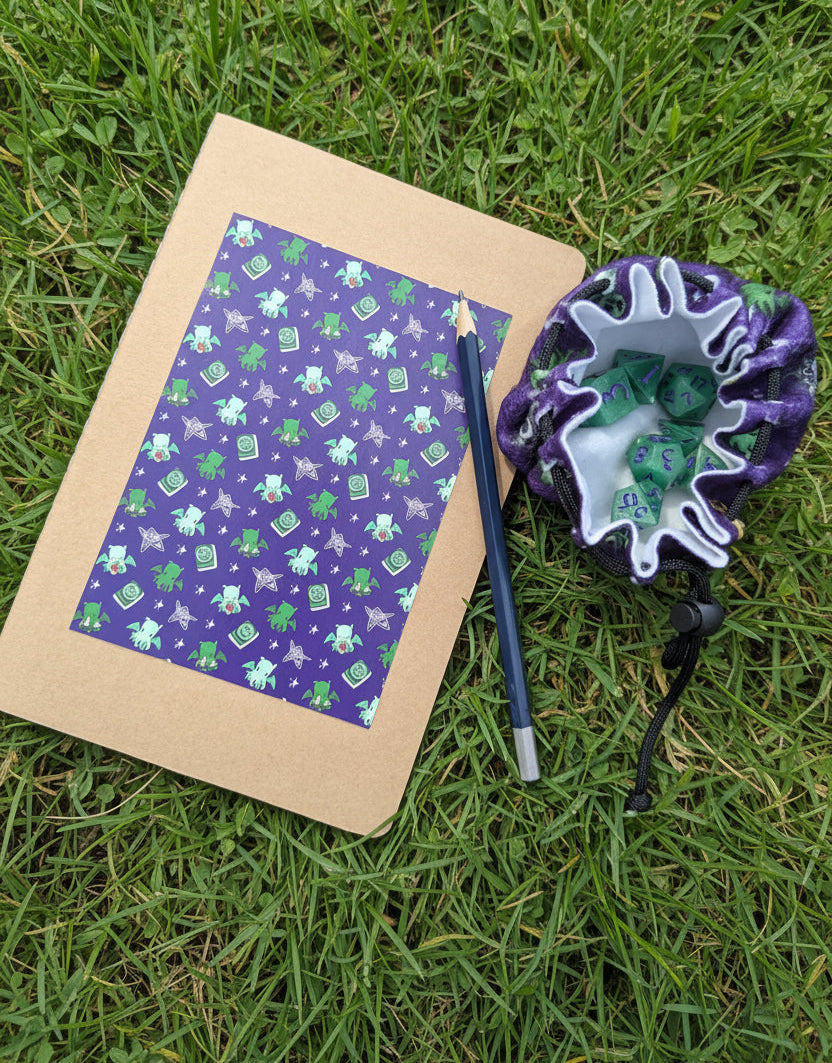 A flat-lay of a kraft paper notebook with a purple and green pattern cover of cthulhu gods, a dark blue pencil, and a matching purple and green patterned drawstring pouch, all arranged on grass.