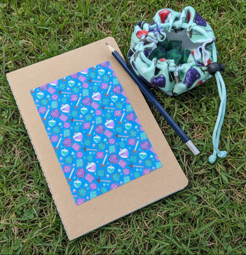 A kraft paper notebook with a colorful patterned cover lies on green grass next to a blue pencil and a mint drawstring pouch filled with green dice.